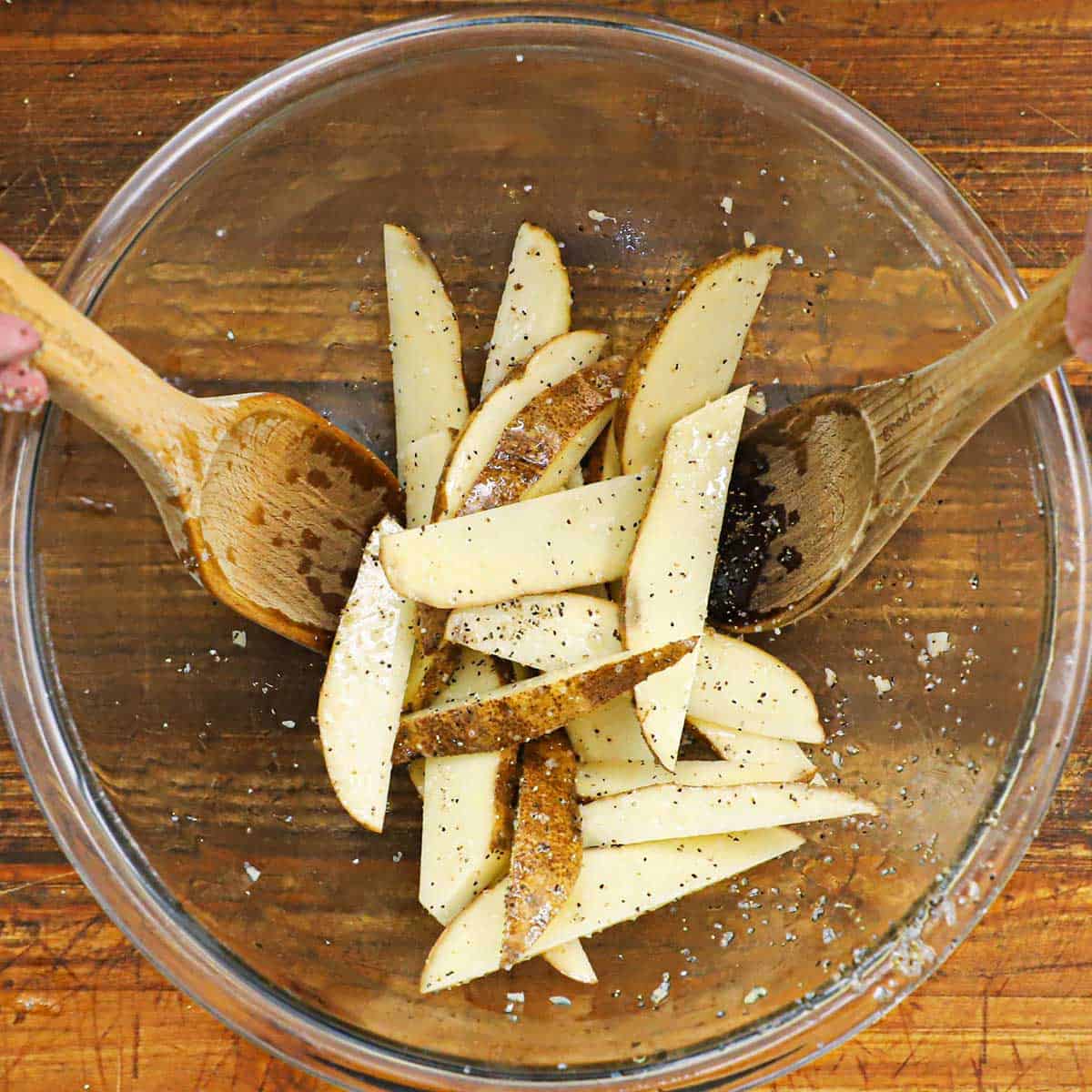 A person using two large wooden spoons to toss potato wedges in a glass bowl with vegetable oil, salt, and pepper.