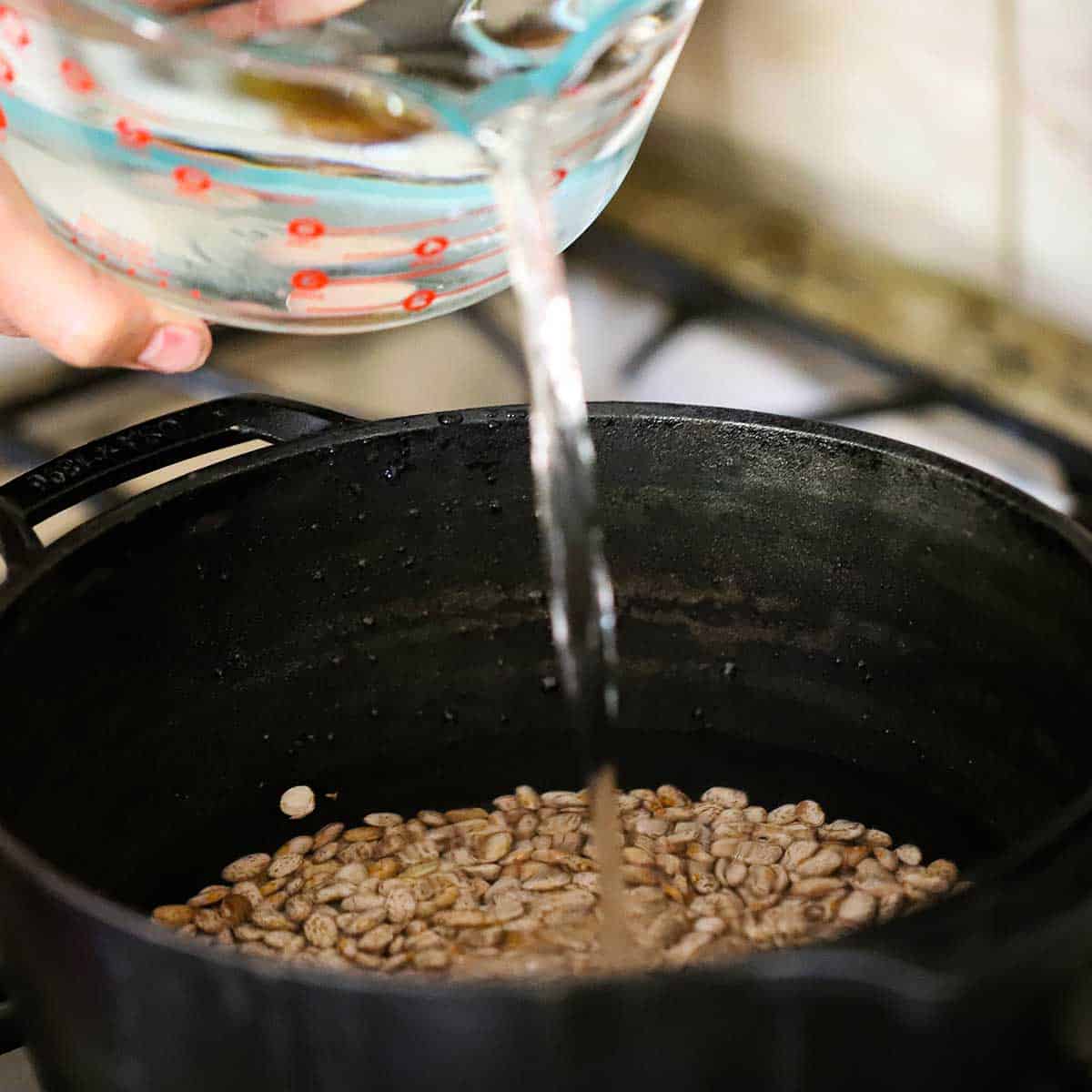 A person pouring water from a large glass measuring cup into a black pot filled with dried pinto beans.