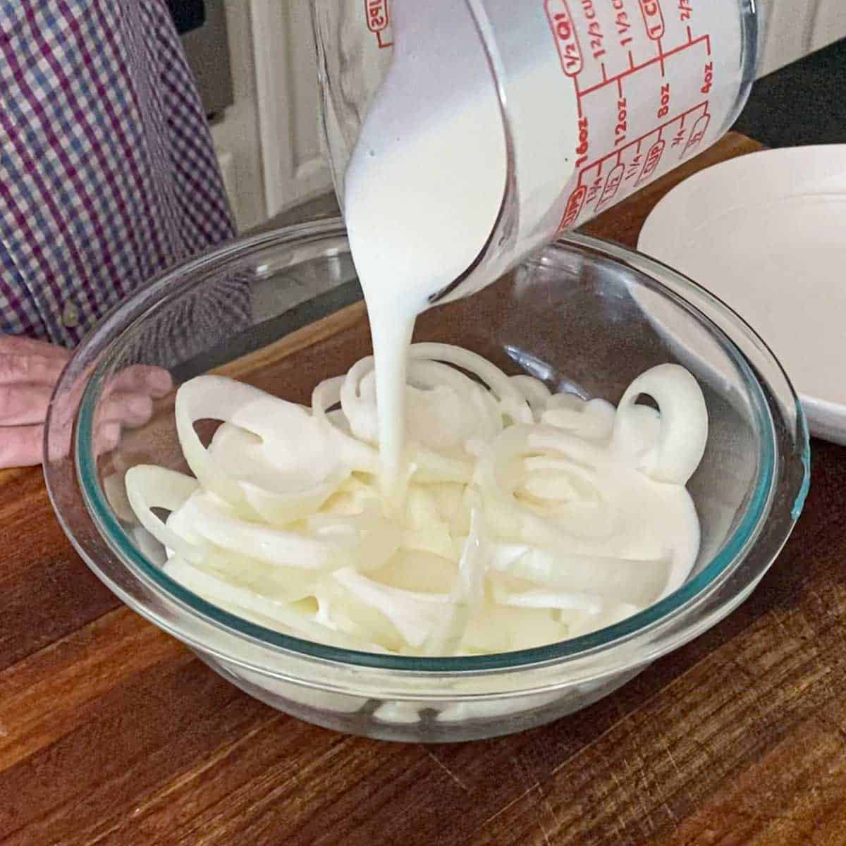 A person pouring buttermilk from a measuring cup over sliced onions in a large glass bowl.