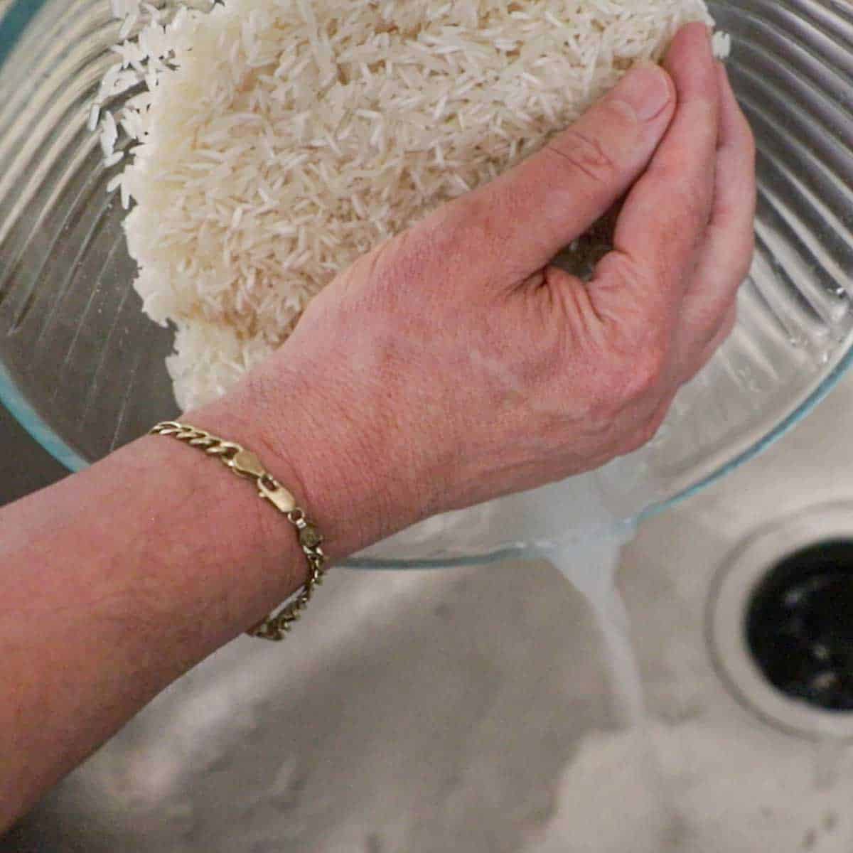 A person using his hand to hold back Jasmine rice that has been rinsed with water in a glass bowl while the water drains into a kitchen sink.