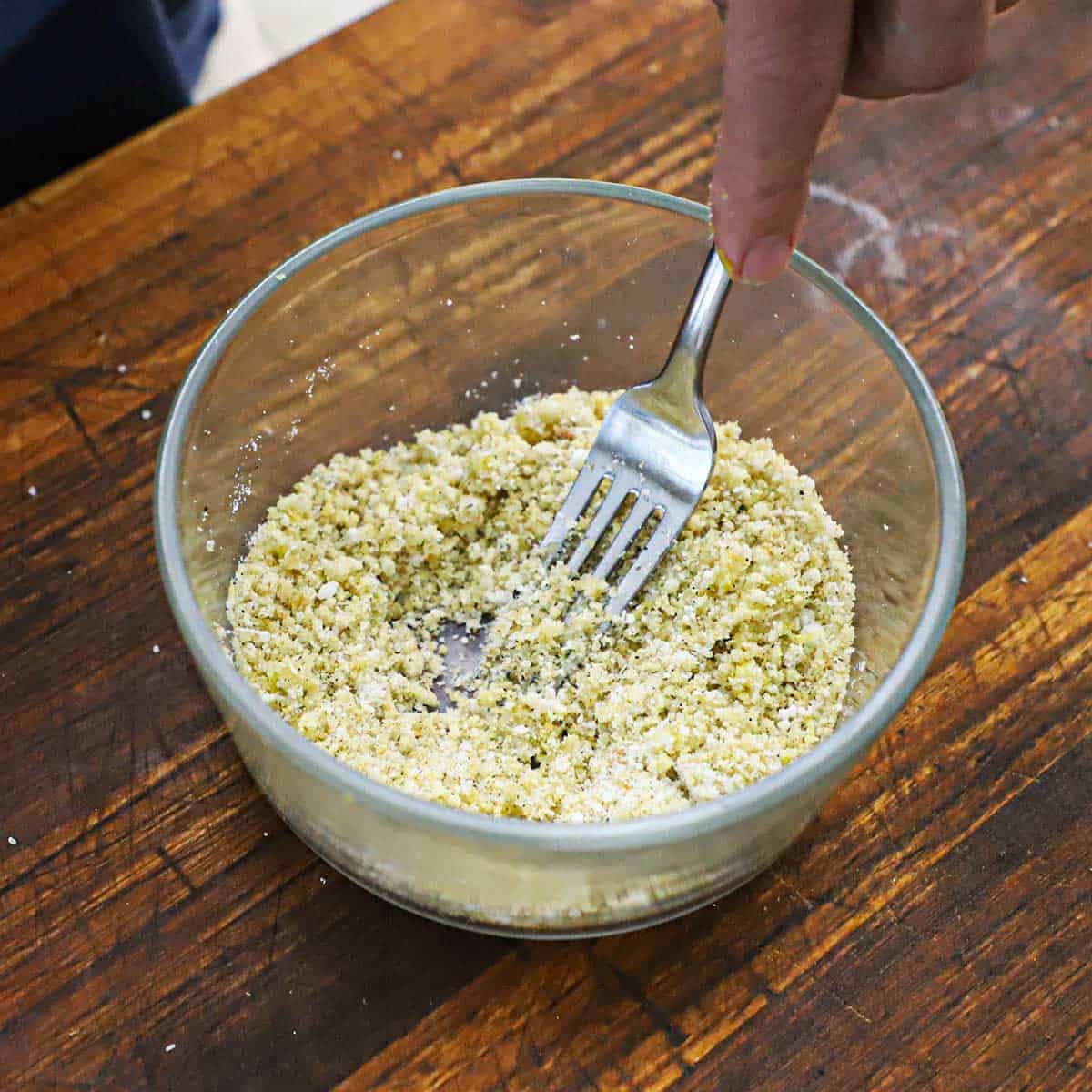 A person using a fork to combine a Panko breadcrumb, herbs, and butter mixture in a small glass bowl.