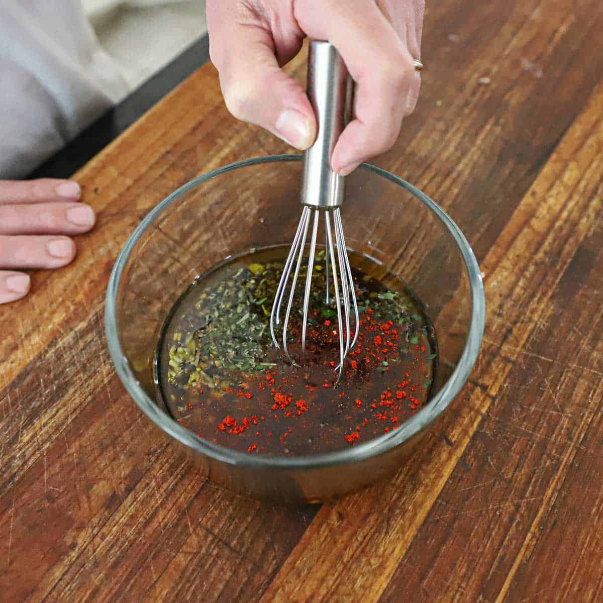 A person using a small whisk to combine the ingredients for a balsamic garlic marinade for steak kabobs in glass bowl on a wooden cutting board.