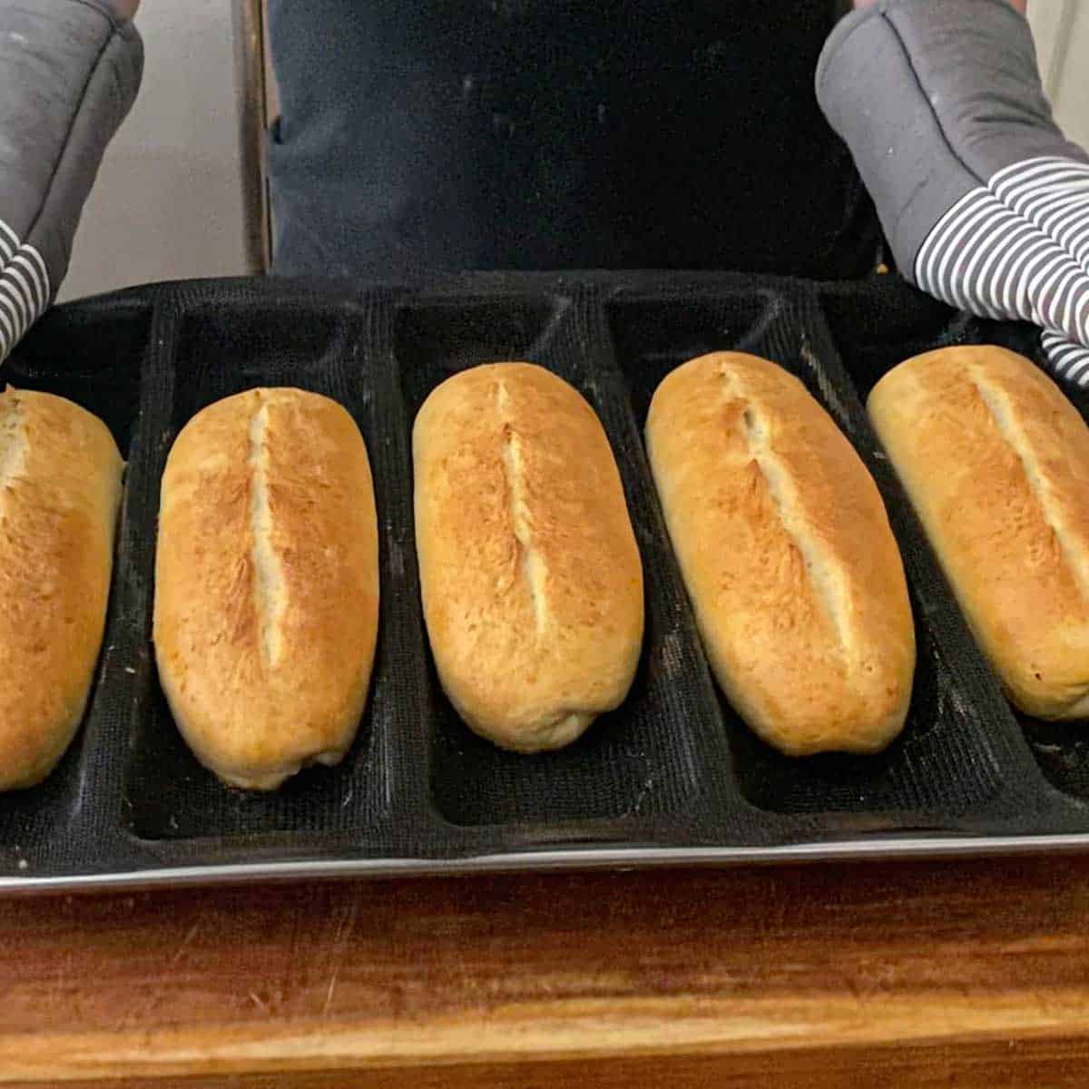 A person holding a pan that is holding an aerated silicone mat with channels containing freshly baked homemade Italian rolls on a wooden cutting board.