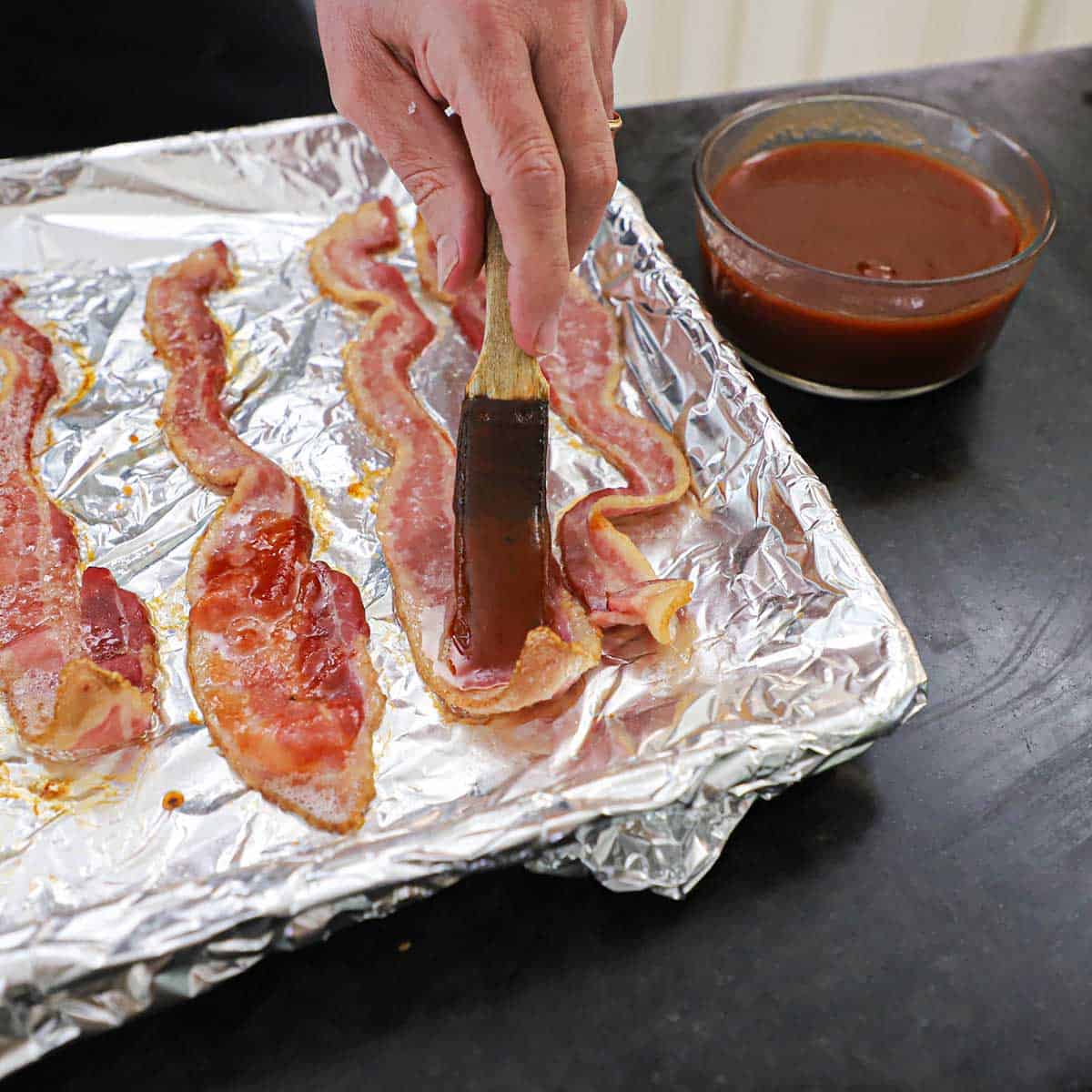 A person using a brush to apply homemade BBQ sauce to partially cooked thick strips of bacon on a foil-lined baking pan.
