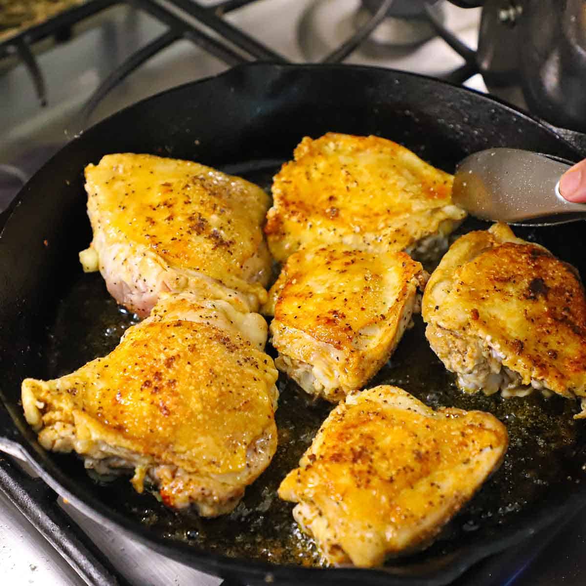 A person using a pair of metal tongs to turn skin-on chicken thighs the are being seared in oil in a large cast-iron skillet.