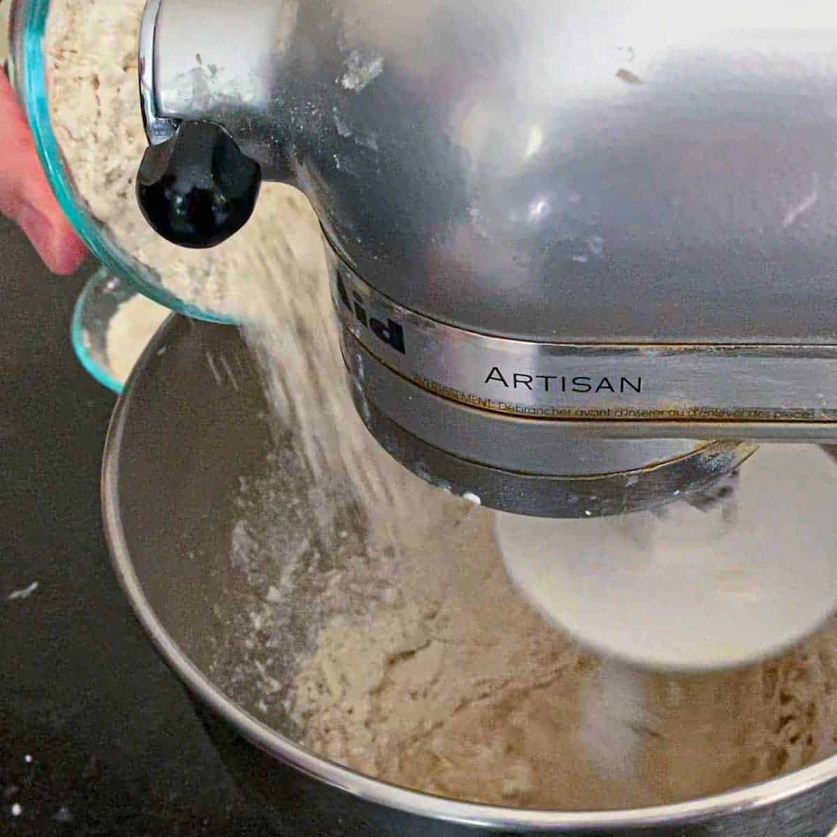 A person dumping bread flour from a small bowl into the bowl of a stand mixer that is mixing Italian roll dough.