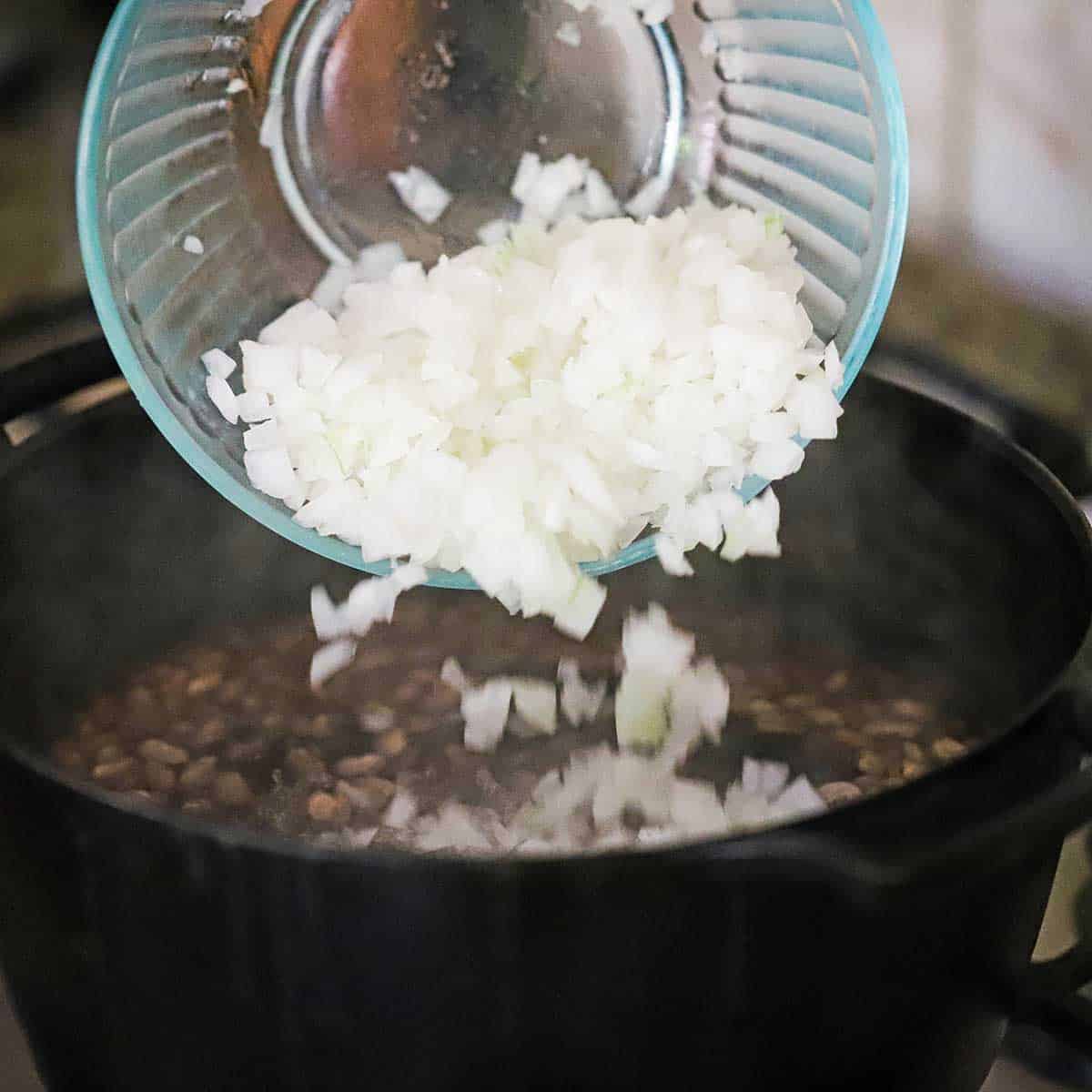 A person dumping chopped onions from a glass bowl into a pot filled with simmering pinto beans in water.