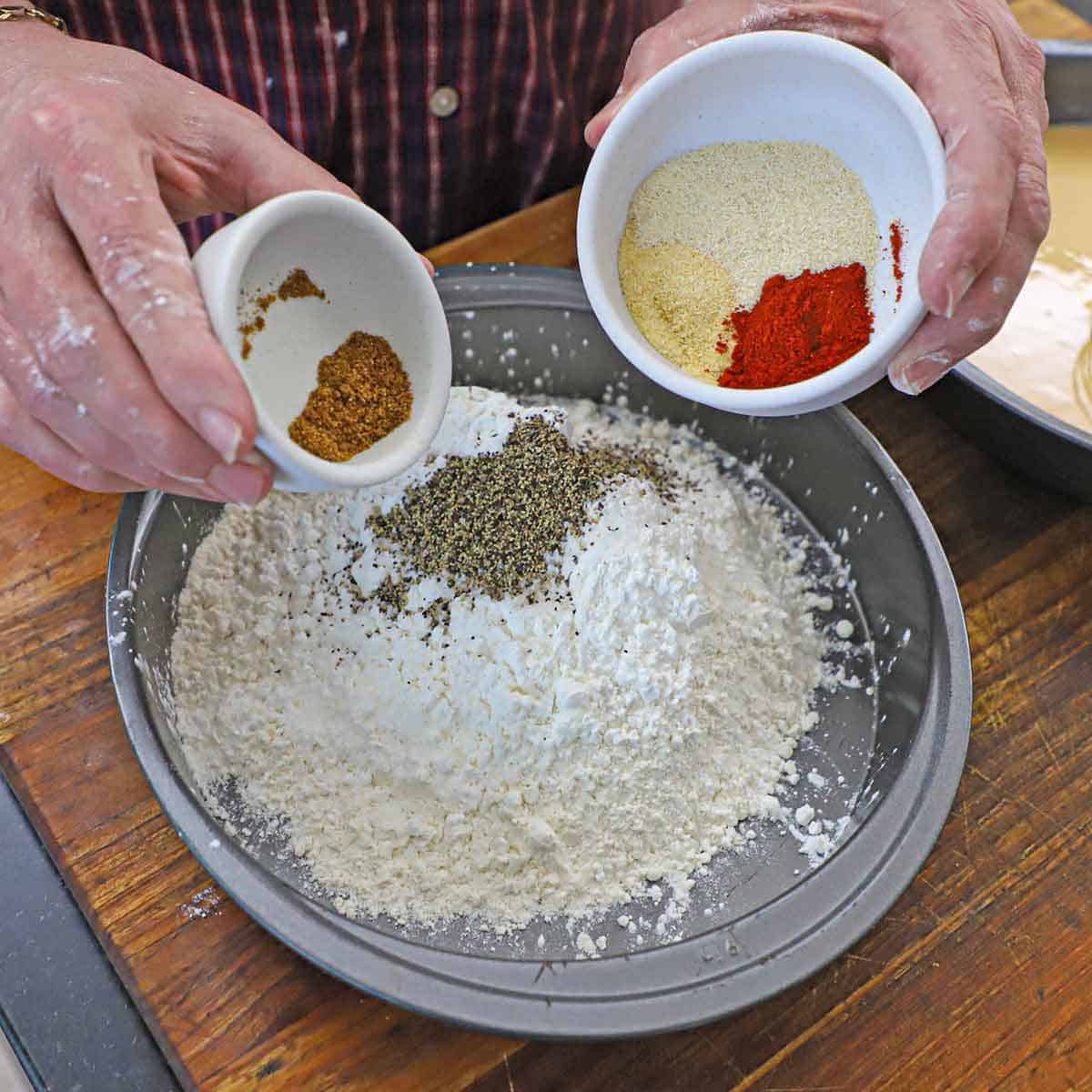 A person holding a small white bowl with cayenne pepper in it in one hand and a small white bowl with more seasonings in his other hand both over a baking pan filled with flour and black pepper.
