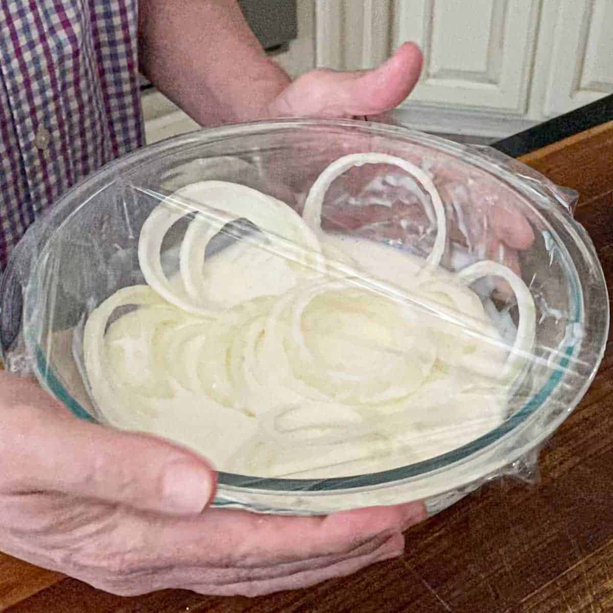 A person holding a glass bowl that is filled with sliced onions covered in a buttermilk with a layer of plastic wrap over the top of the bowl.