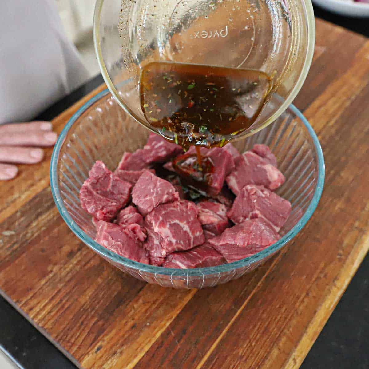 A person pouring a garlic balsamic marinade from a small glass bowl into another glass bowl that is filled with large chunks of beef tenderloin.