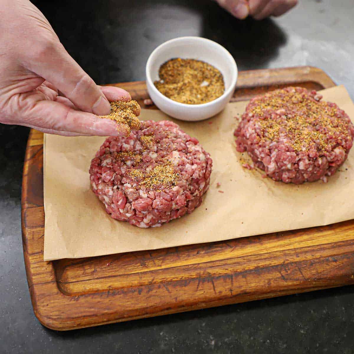A person sprinkling BBQ rub over the tops of two raw sirloin burger patties that are resting on brown paper on a small wooden cutting board.