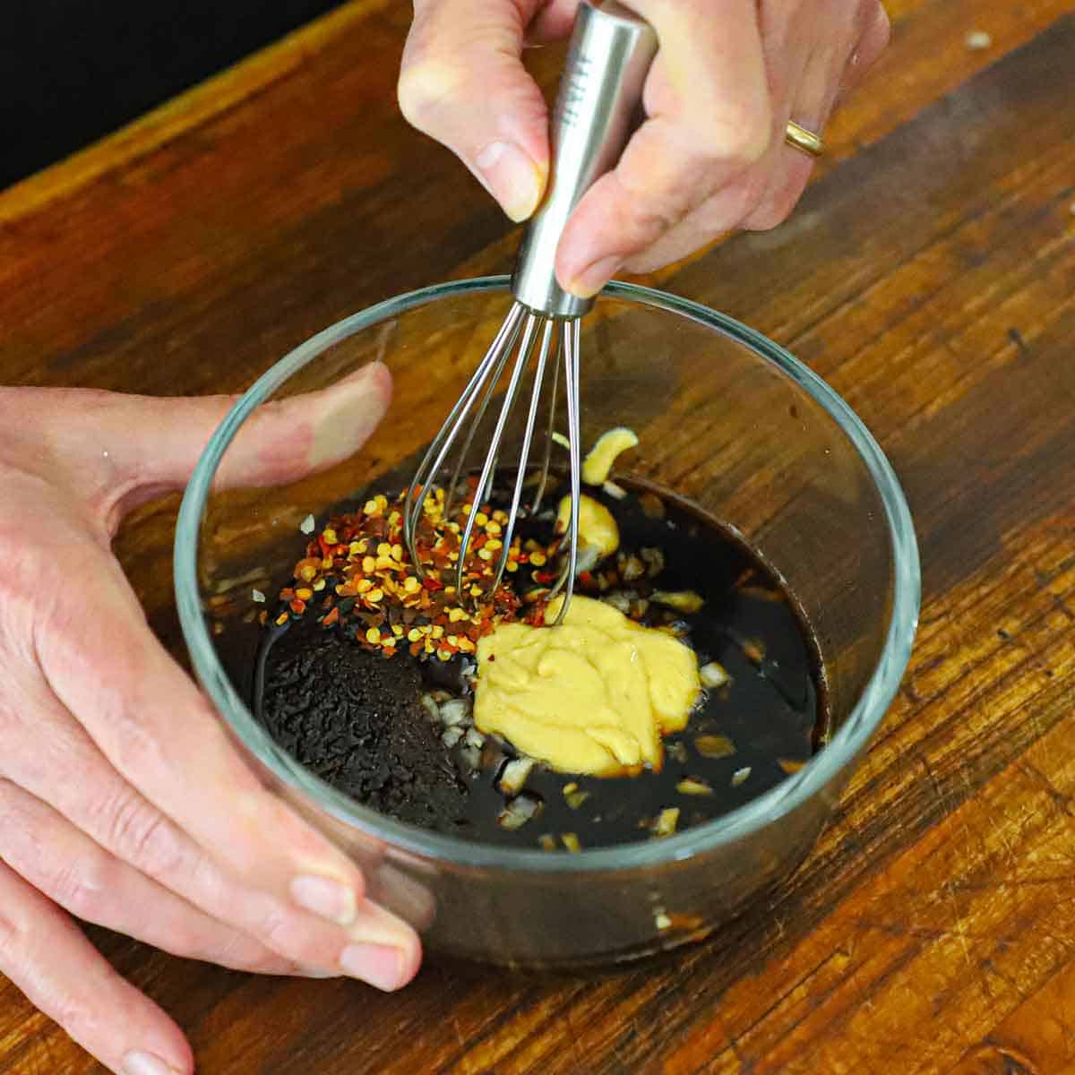 A person using a small whisk to combine brown sugar, soy sauce, vinegar, mustard, and red pepper flakes in a small glass bowl.