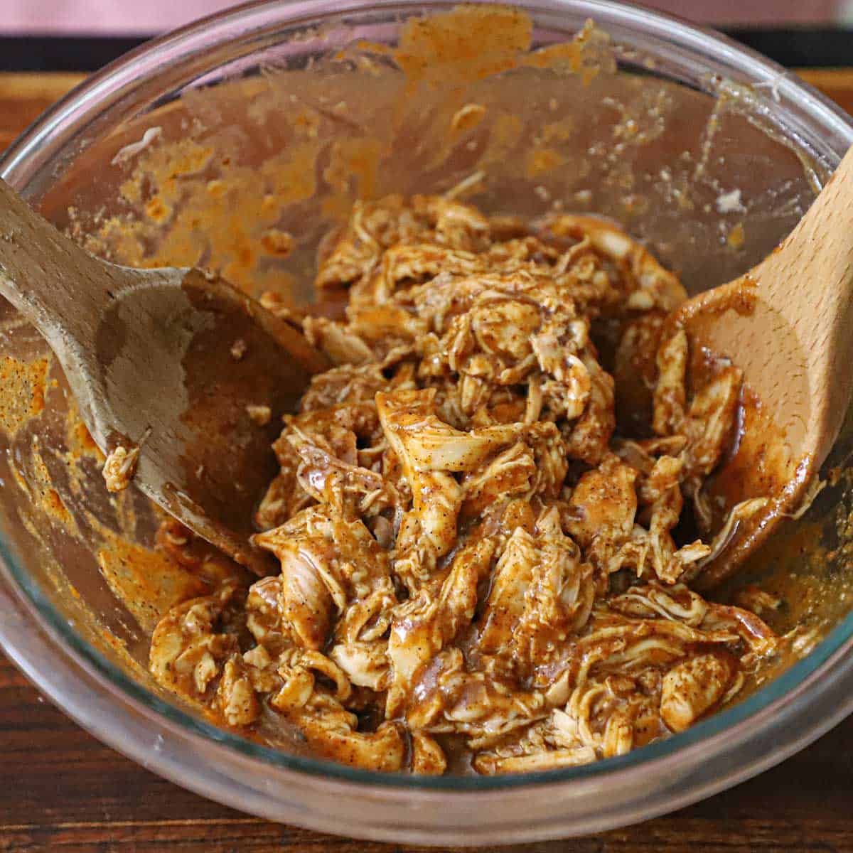 A person using two wooden spoons to toss shredded chicken in a red enchilada sauce in a large glass bowl.