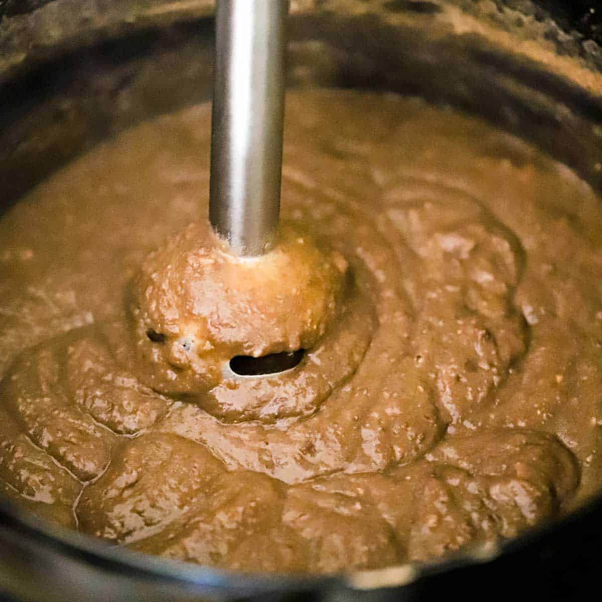 A person using an immersion blender to purée pinto beans that have been softened in simmering water on the stove.