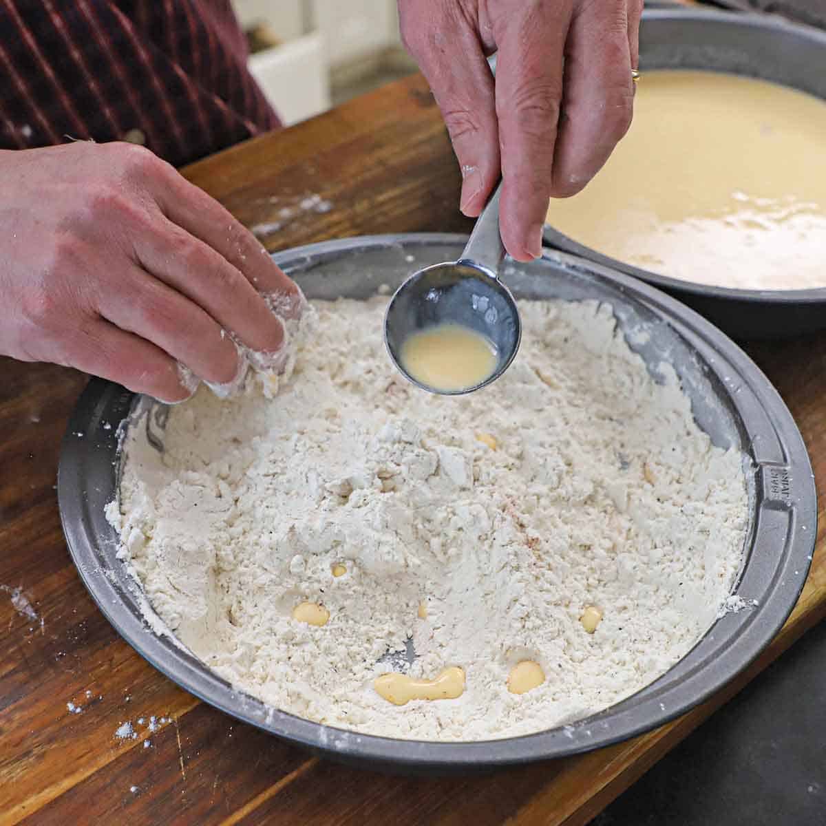 A person using a tablespoon to drizzle wet fried chicken batter into a baking pan filled with seasoned flour.