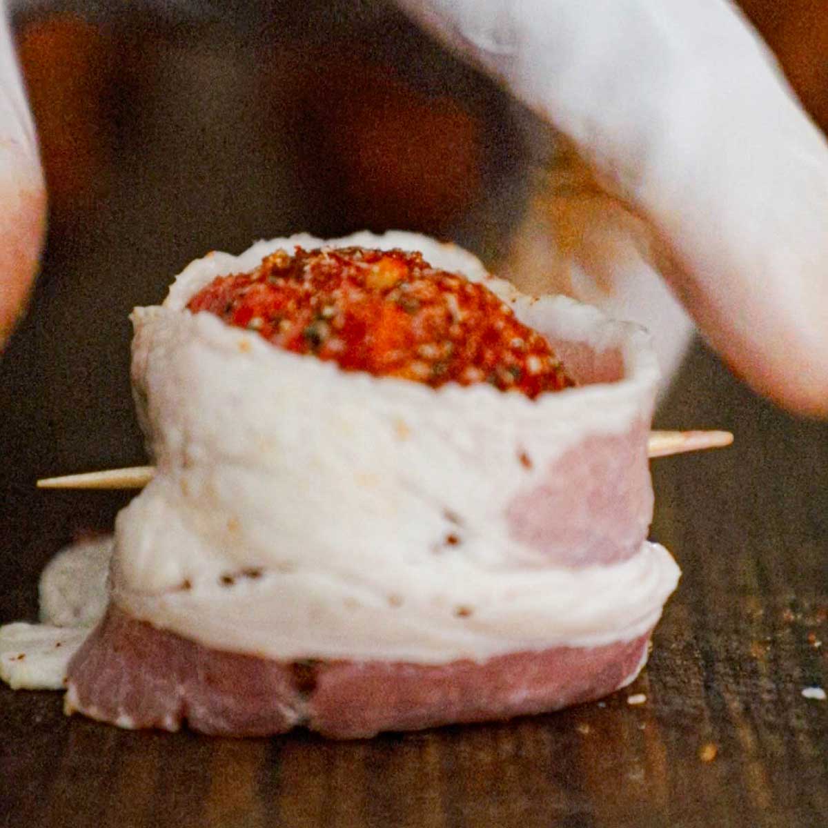 A person placing a toothpick through an uncooked BBQ seasoned meatball that is wrapped with a thin strip of bacon.