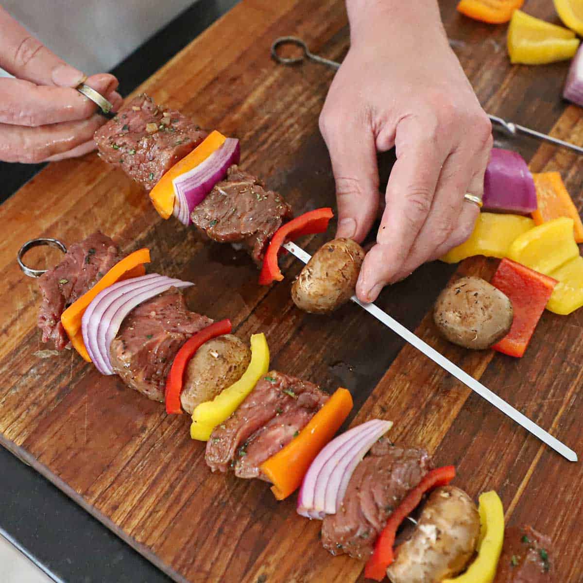 A person using his hand to skewer vegetables, mushrooms, and chunks of marinated beef tenderloin onto metal skewers on a wooden cutting board.