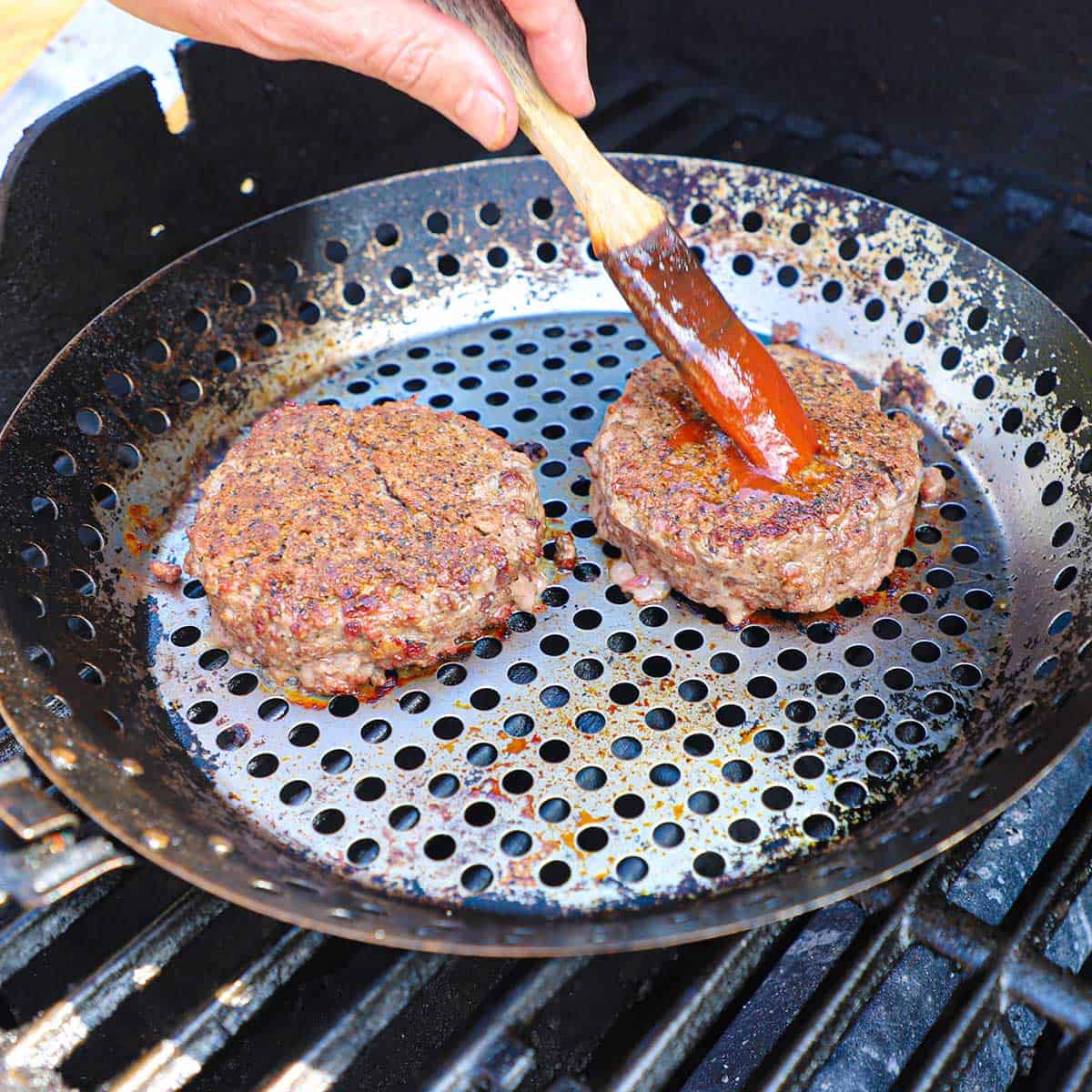 A person using a brush to apply BBQ sauce onto two burgers that are being cooked in a grill pan on a gas grill.