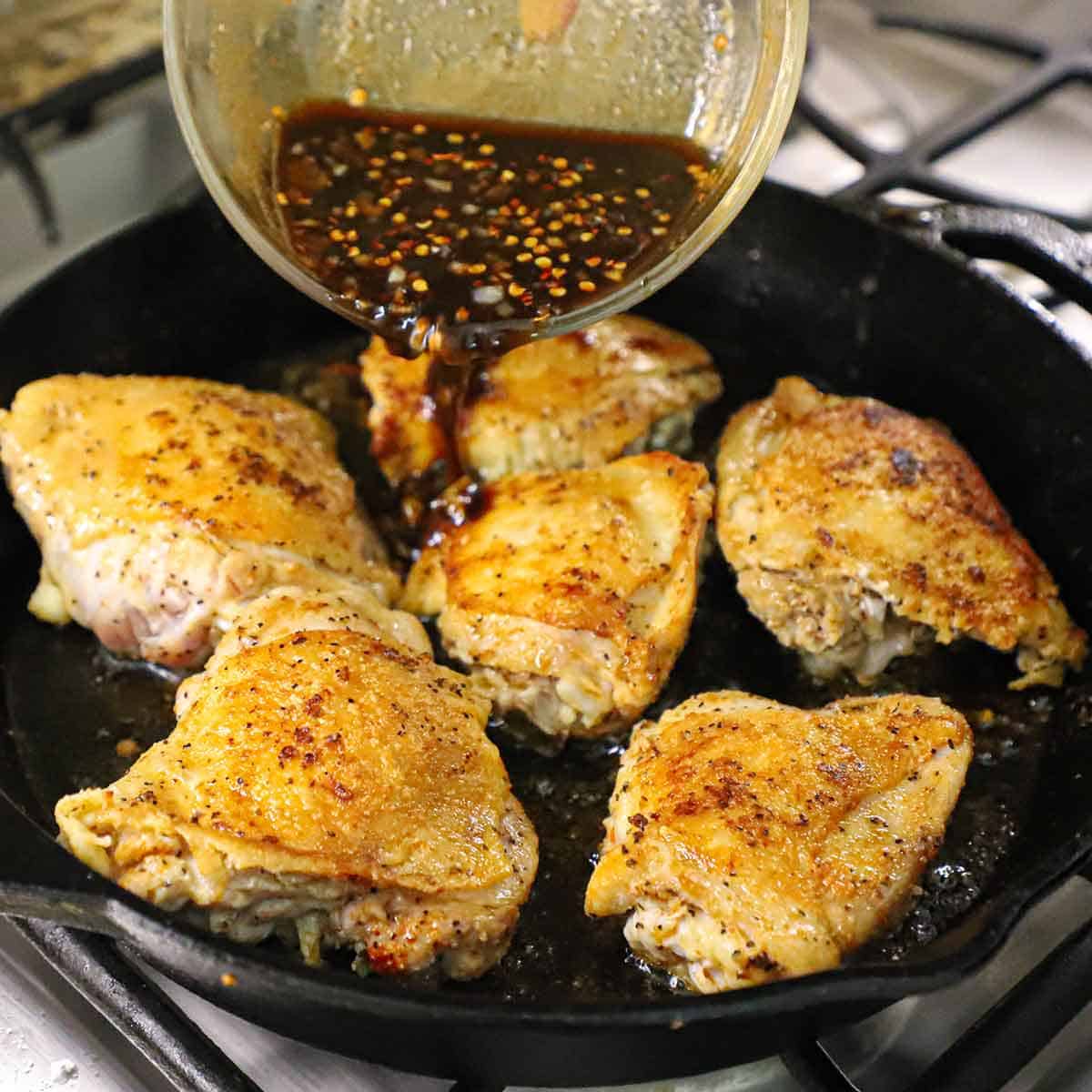 A person pouring a brown sugar garlic sauce around chicken thighs that have been seared in a cast-iron skillet and are still in the skillet.