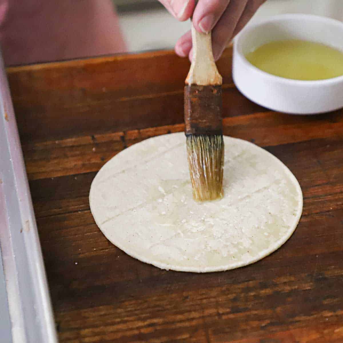 A person using a pastry brush to apply a thin layer of vegetable oil over the surface of a white corn tortilla.