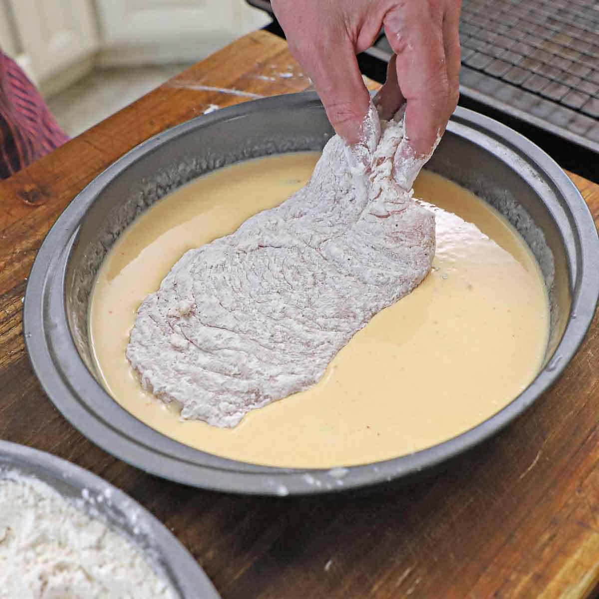 A person dredging a thin floured chicken cutlet through a wet fried chicken batter in a round baking pan.