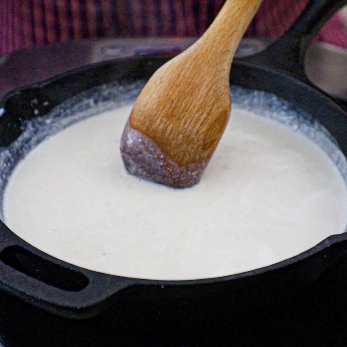 A person using a wooden spoon to stir cream gravy that is just starting to thicken in a cast-iron skillet.