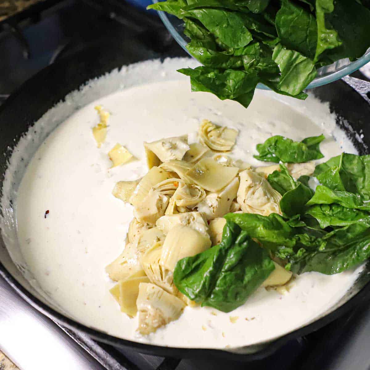 A person transferring chopped fresh spinach leaves into a cast-iron skillet filled with a thick, simmering cream sauce and artichoke hearts.