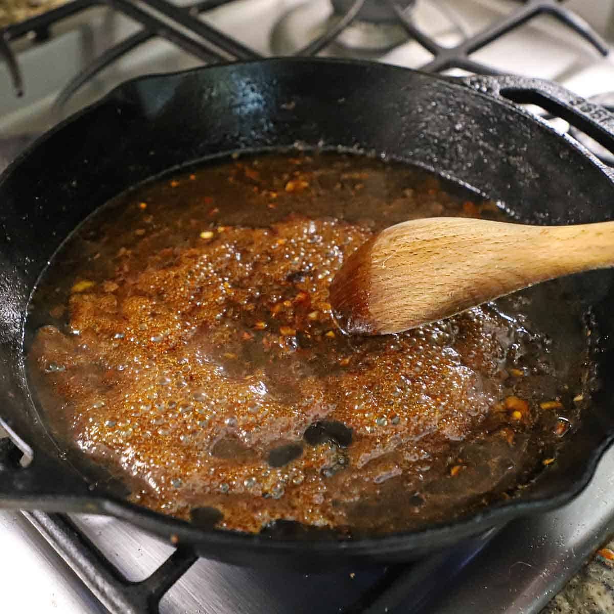 A person using a wooden spoon to stir a simmering brown sugar garlic glaze in a cast-iron skillet on a gas stove.