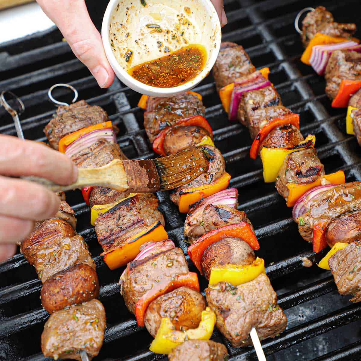 A person using a brush to apply a marinade over the tops of steak kabobs that are being grilled on a gas stove.