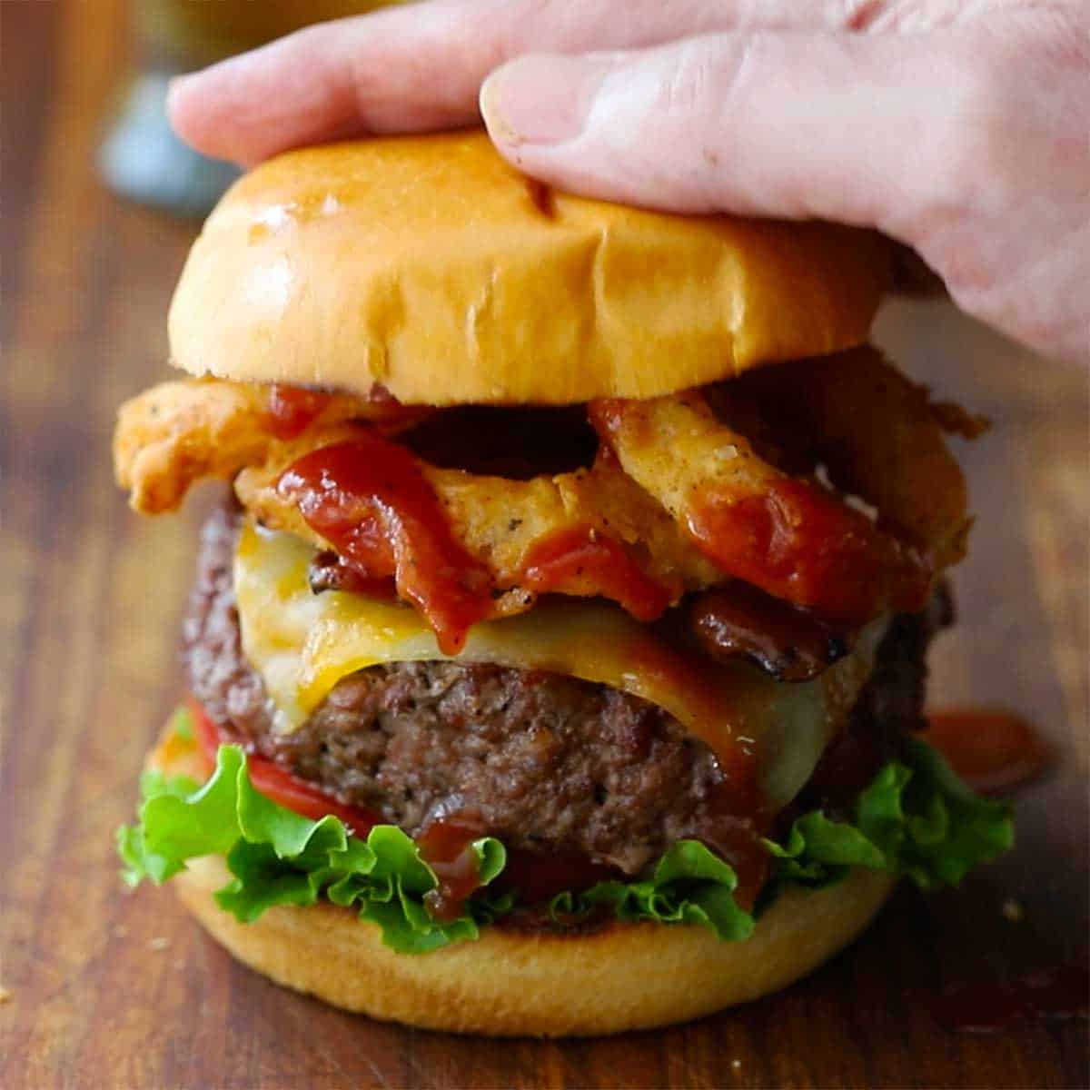 A person pressing down on the top of Brioche bun that is resting on the top of BBQ Bacon Burger with cheese and onion rings.