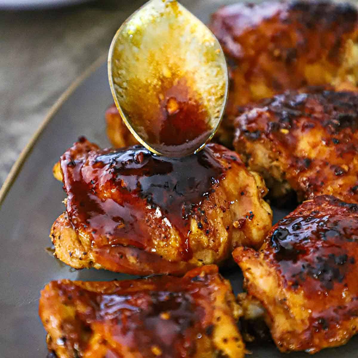 A person using a serving spoon to pour a brown sugar garlic glaze over the top of a cooked skin-on chicken thigh on a platter next to more cooked chicken thighs.