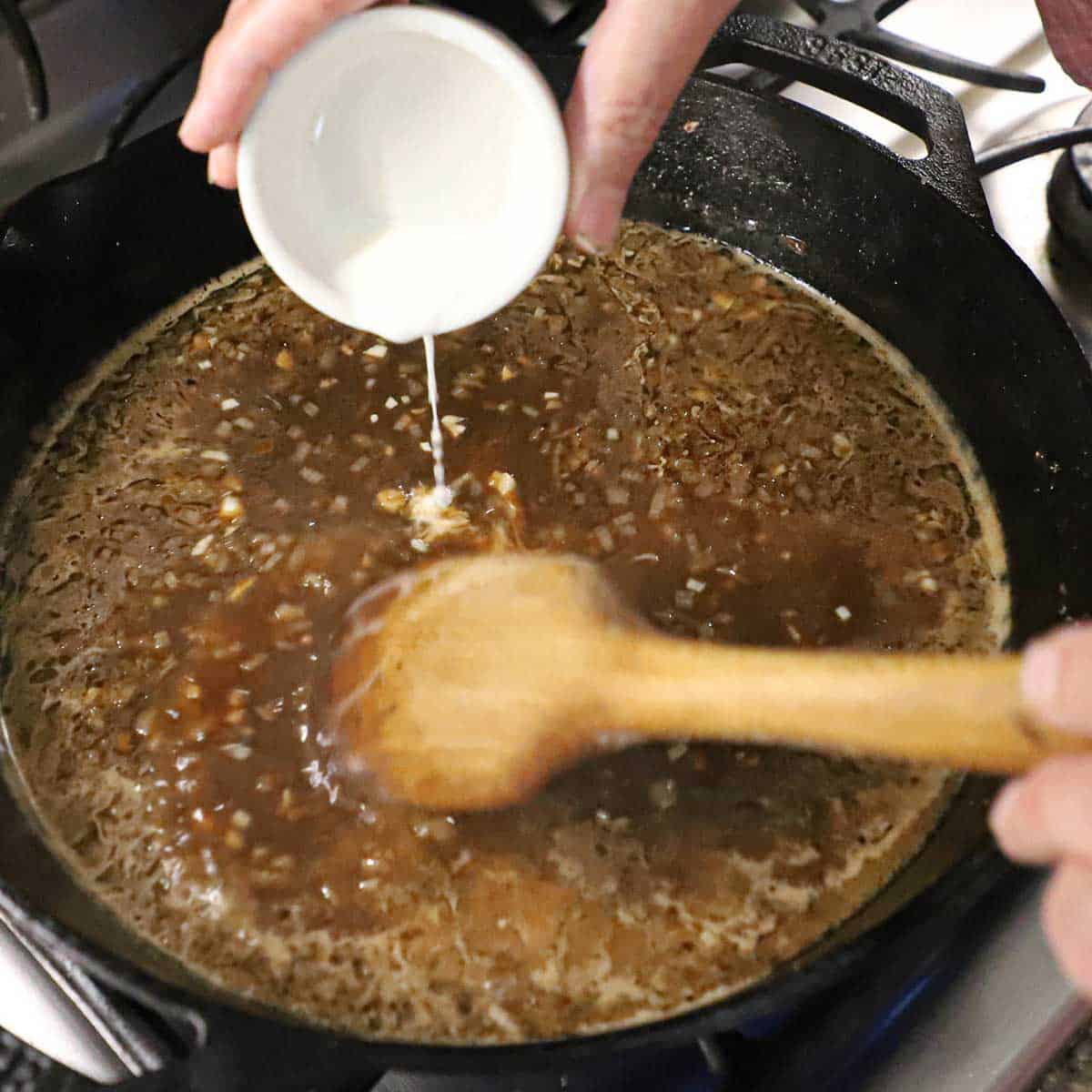 A person pouring a cornstarch slurry from a small white bowl into a skillet that is filled with a beef stock and Madeira wine sauce.