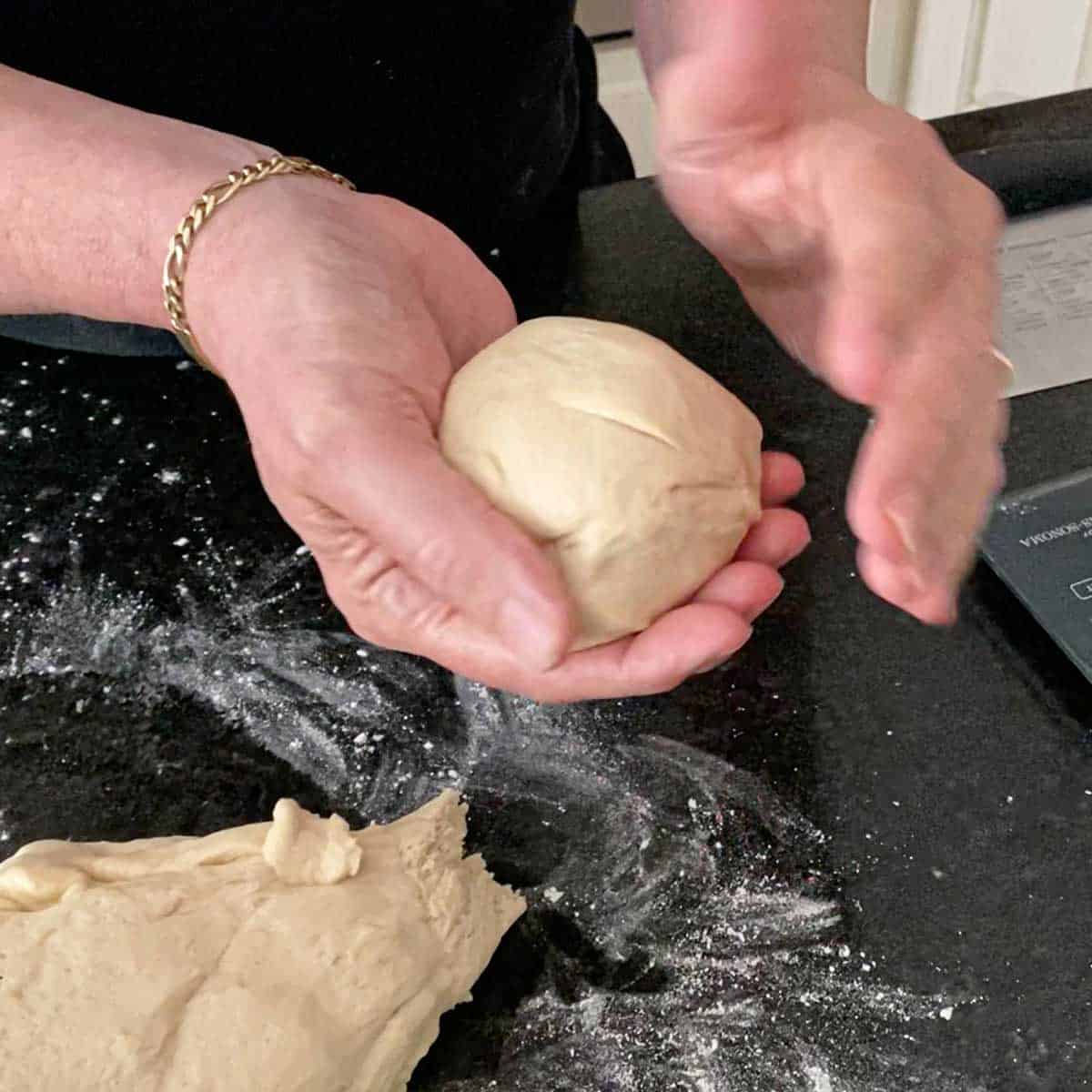 A person forming a ball of dough in his hands that he has pinched for a large ball of dough on a lightly floured marble counter.