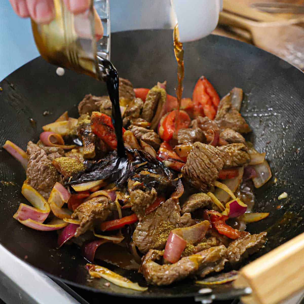A person pouring Peruvian soy sauce from one bowl and red wine vinegar from another bowl into a wok that is filled with pieces of beef tenderloin and vegetables that are being stir-fried.