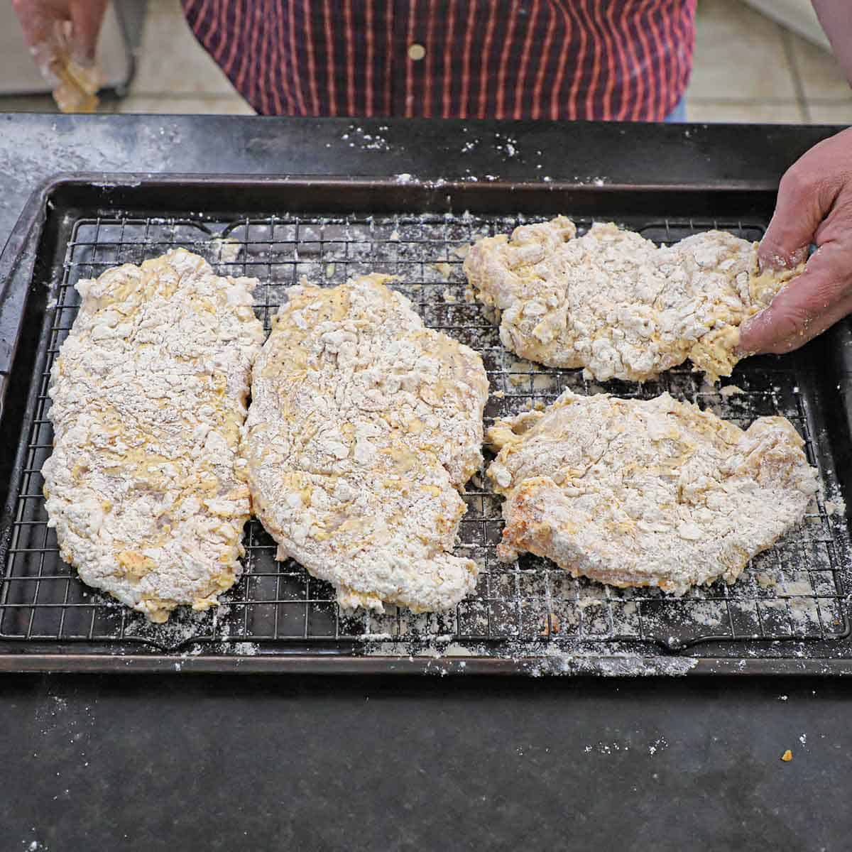 A person placing uncooked bread chicken cutlets on a wire rack in a baking pan that is nestled into a baking sheet pan.