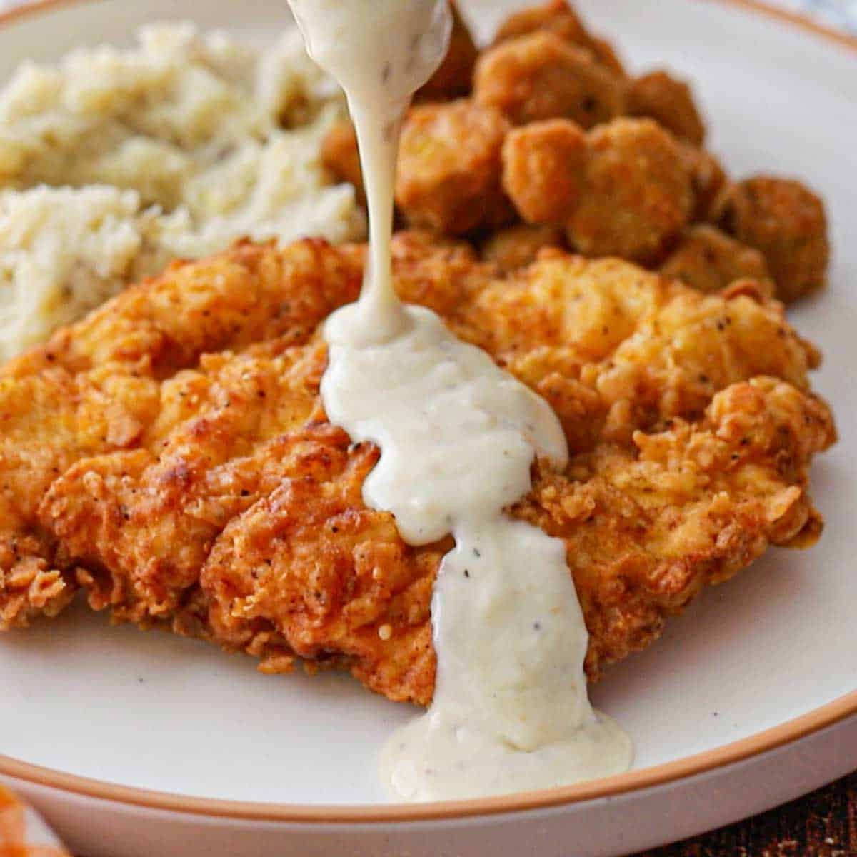 A person pouring Classic Southern Cream Gravy from a glass gravy boat over a cutlet of chicken fried chicken on a white dinner plate.