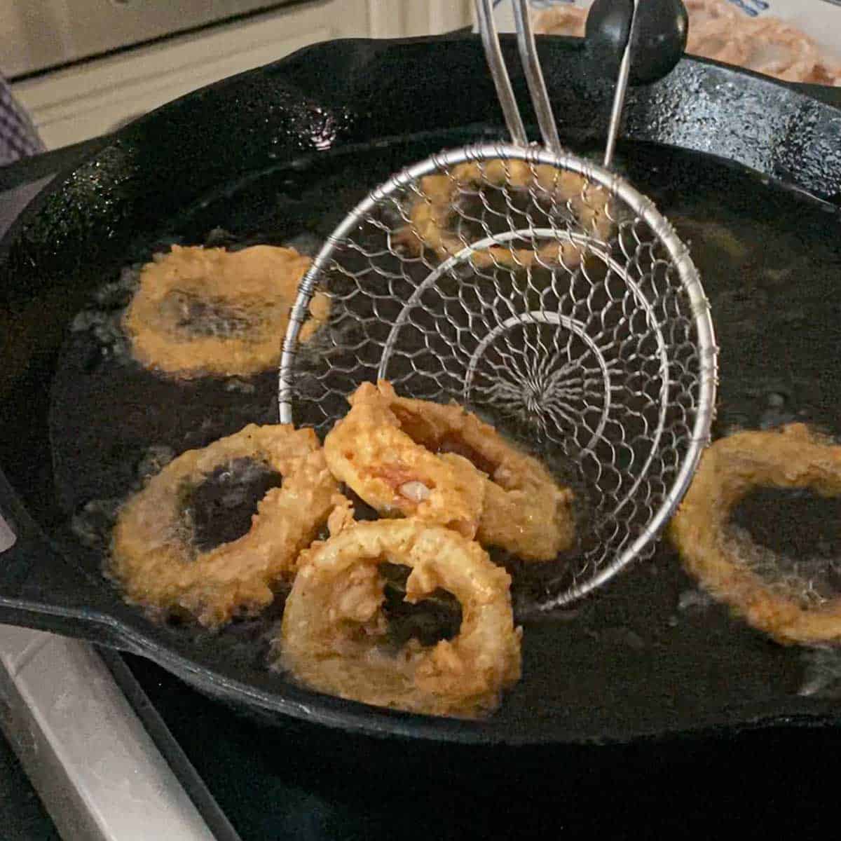 A person using a metal spider to lift crispy steakhouse onion rings out a hot oil in a large cas-iron skillet.