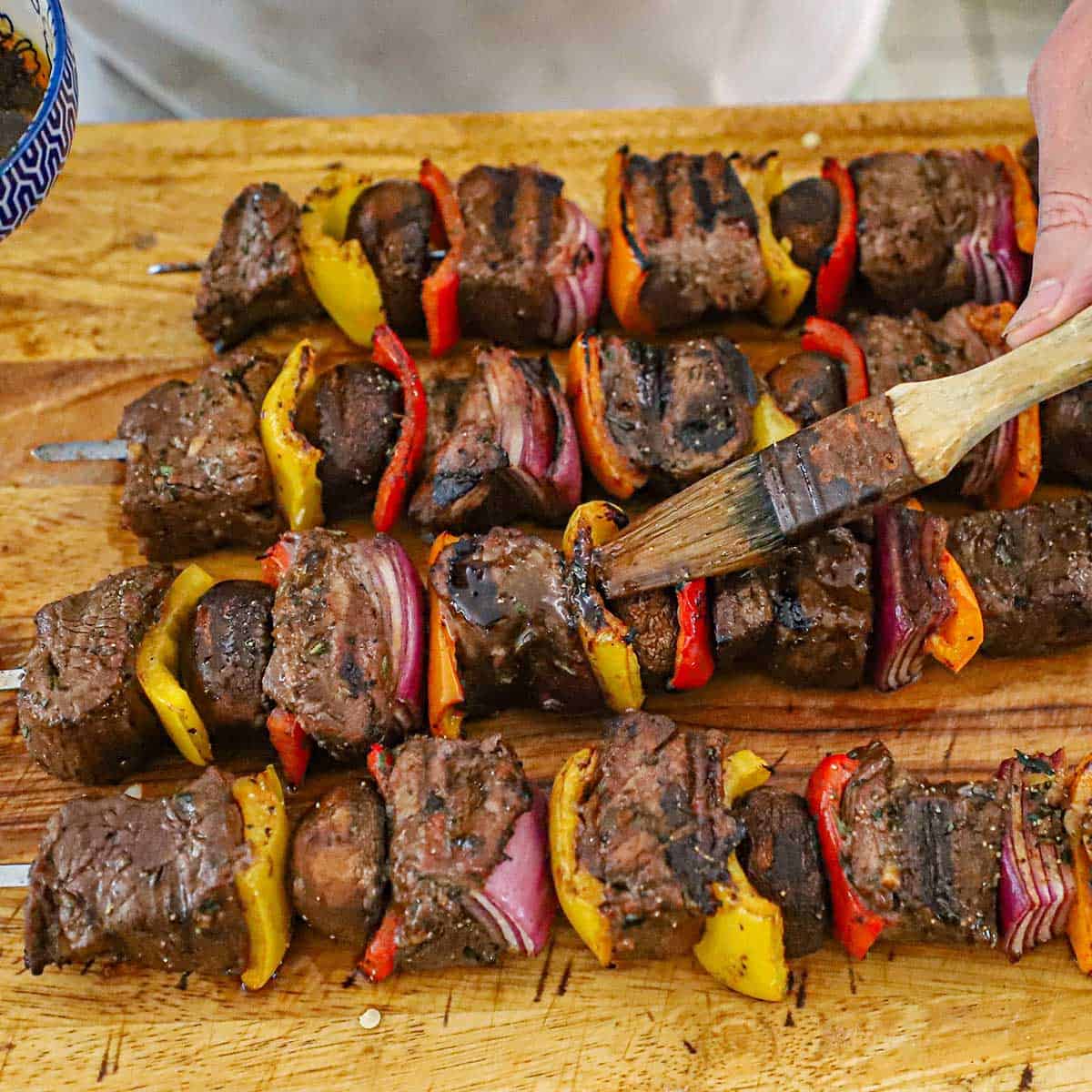 A person using a pastry brush to apply a balsamic sauce over the tops of freshly grilled steak kabobs.