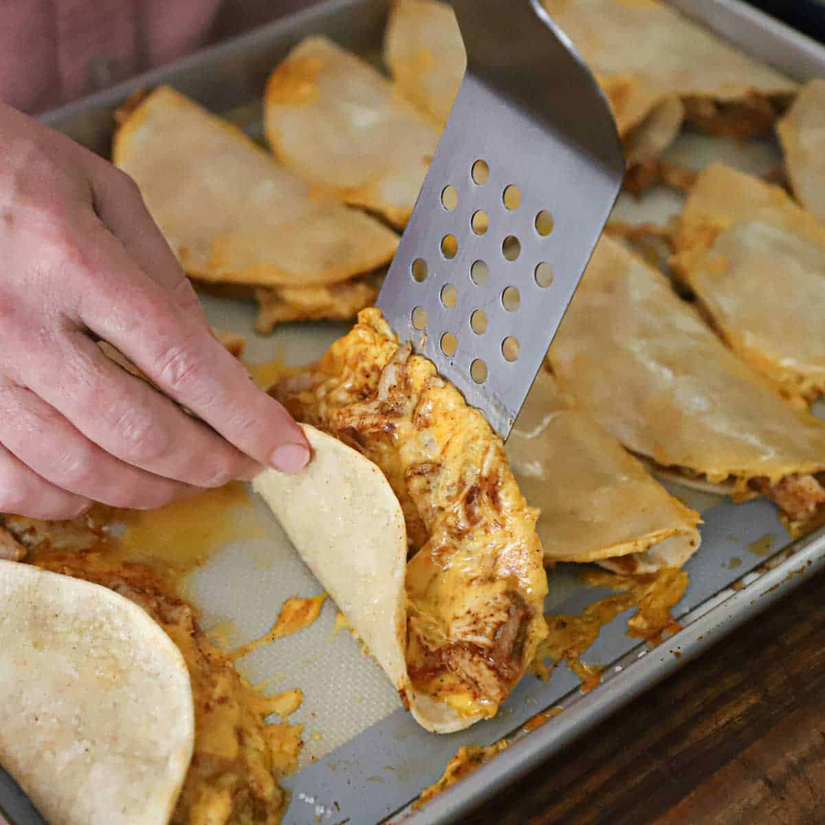 A person using a perforated spatula to flip a baked chicken taco over on a baking sheet lined with a silicone matt and other tacos.