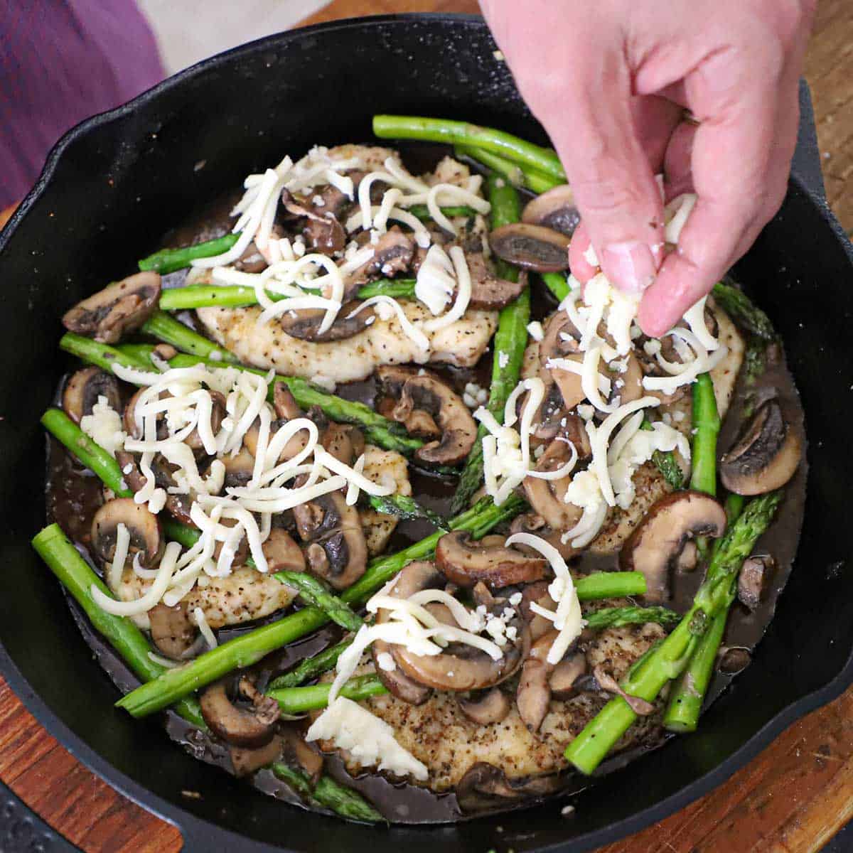 A person sprinkling shredded mozzarella cheese over the top of chicken Madeira and sautéed mushrooms and asparagus in a cast-iron skillet.