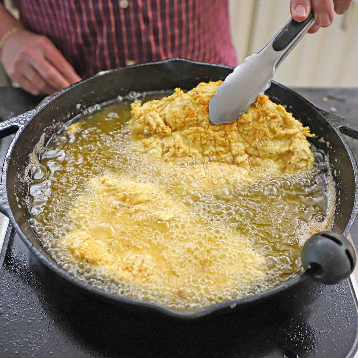 A person using a pair of metal tongs to flip a chicken fried chicken cutlet in a large cast-iron skillet filled with hot, bubbling oil.