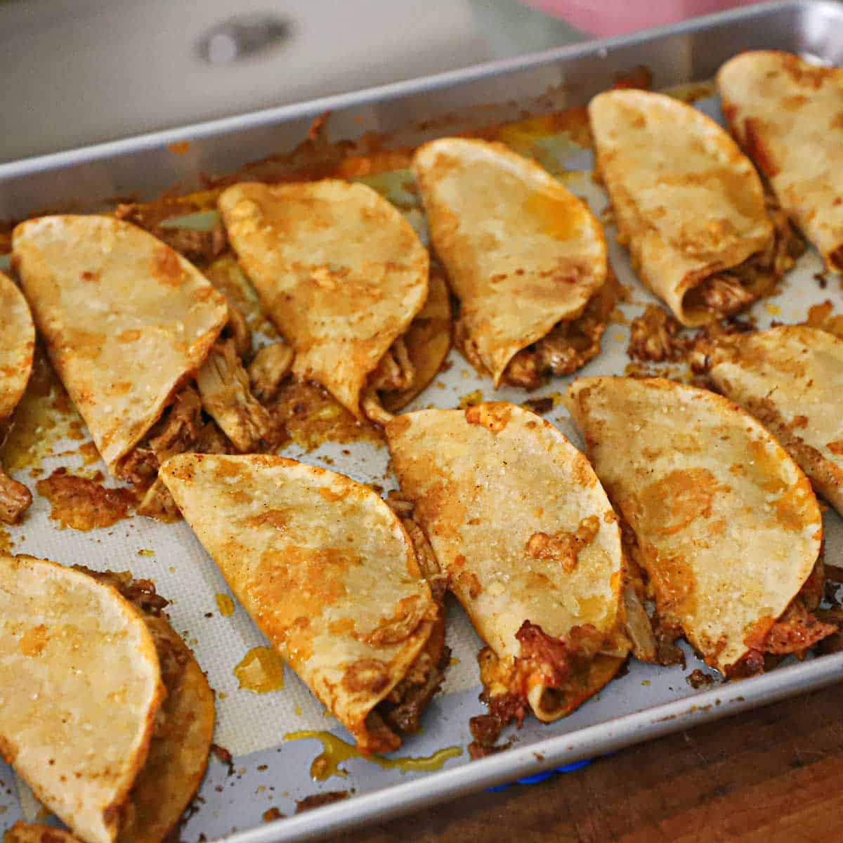 Two rows of crispy baked chicken tacos on a large baking sheet that is lined with a silicone matt.