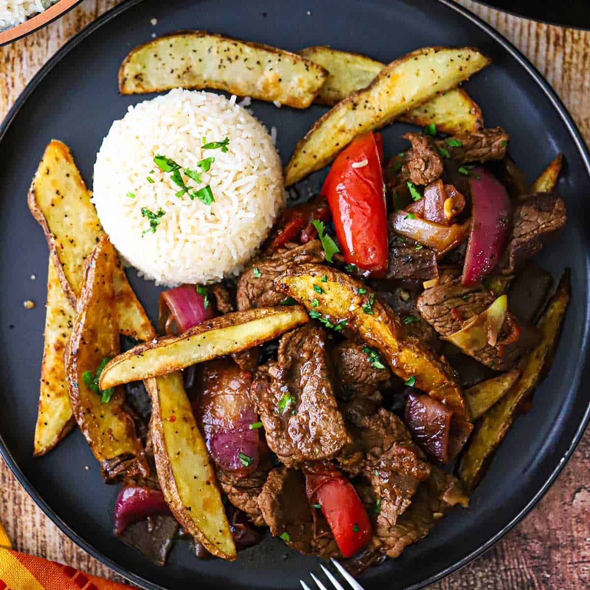 An overhead view of a serving of Classic Lomo Saltado, Peruvian Steak and Potatoes Stir-Fry, on a black dinner plate with a mound of Peruvian Garlic Rice on the plate as well.