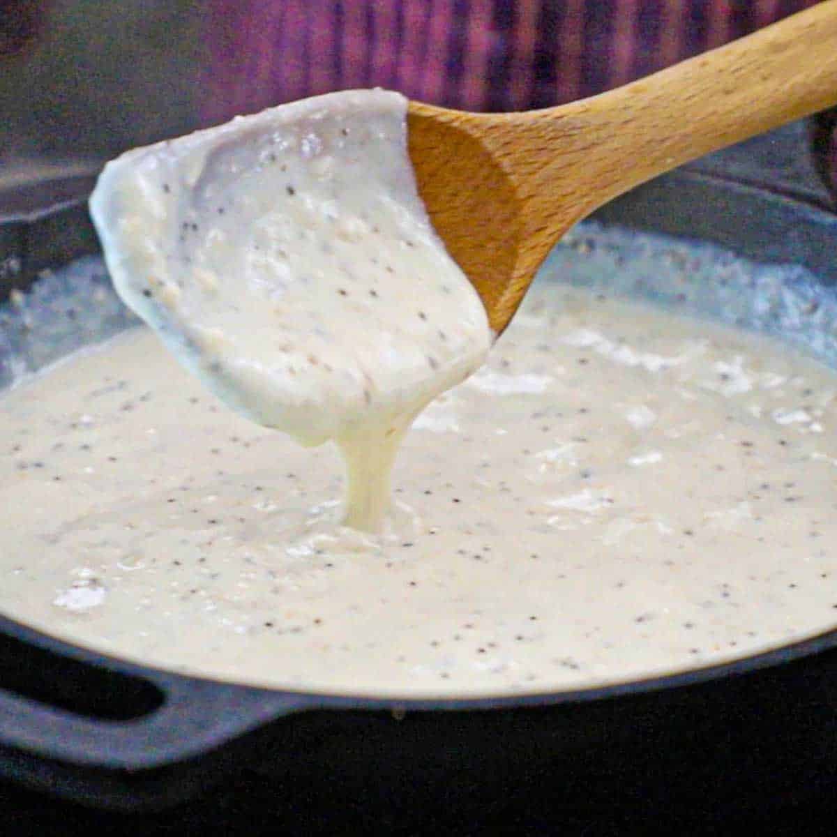 A person lifting a large wooden spoon from a cast-iron skillet that is filled with homemade cream gravy.