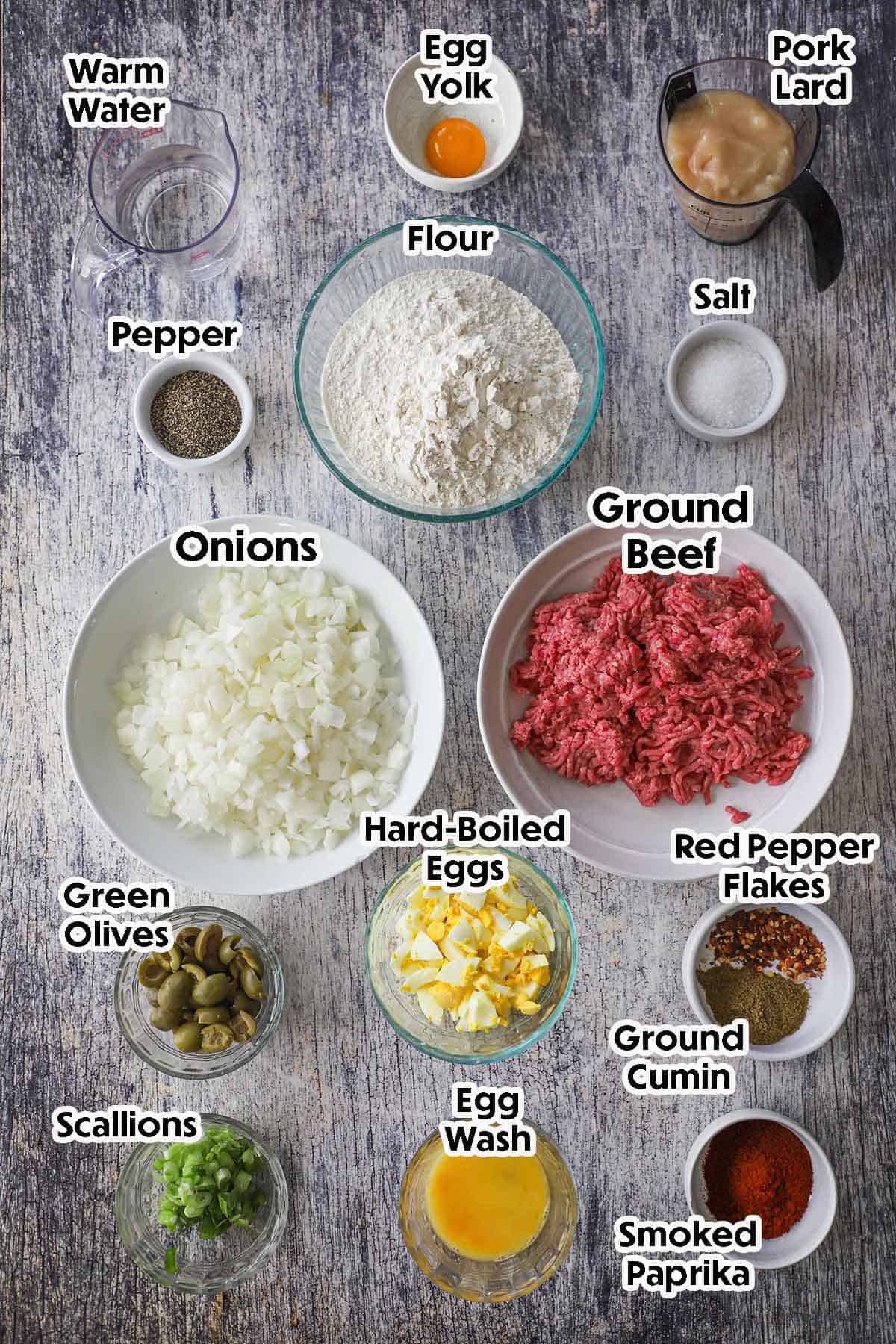 An arrangement of ingredients for Argentine Beef Empanada on a grey wooden background including ground beef, onion, flour, pork lard, hard boiled eggs, olives, and seasonings.