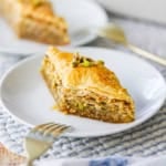 A straight-on view of a diamond-shaped piece of Authentic Greek Baklava on a white dessert plate with a gold fork nearby.