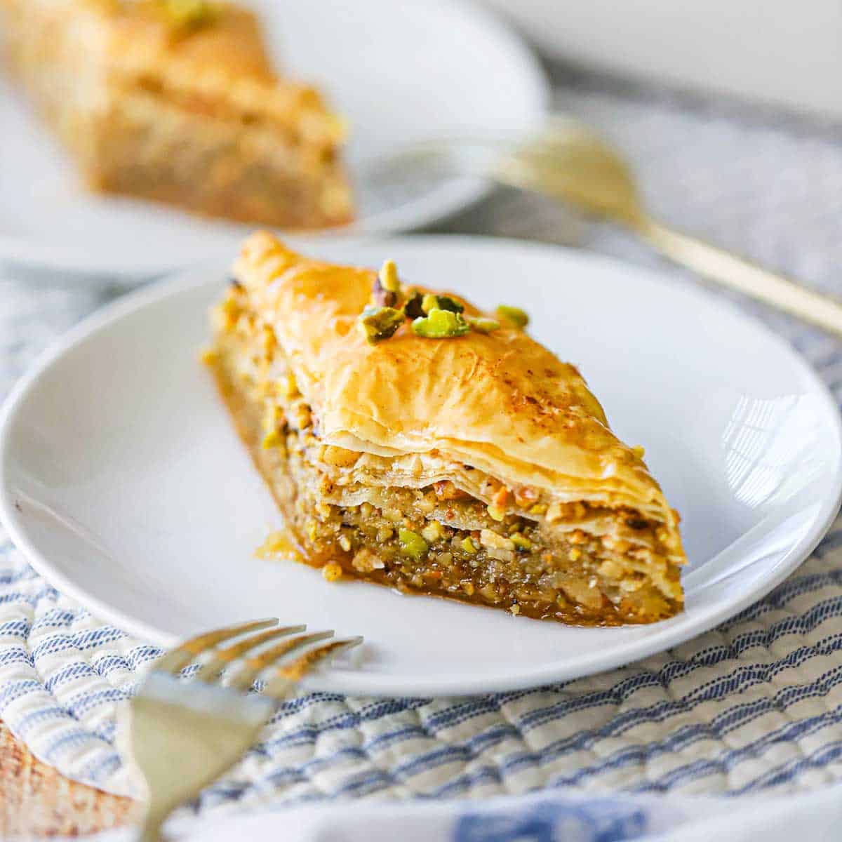A straight-on view of a diamond-shaped piece of Authentic Greek Baklava on a white dessert plate with a gold fork nearby.
