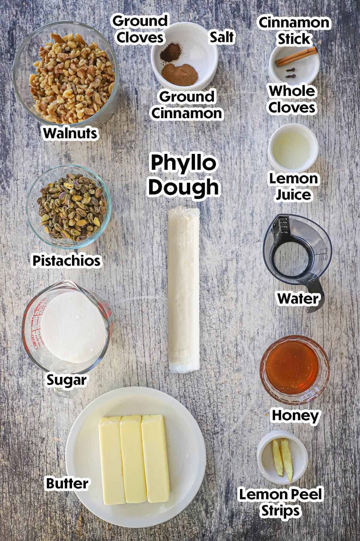 An arrangement of ingredients for Authentic Greek Baklava on a grey wooden background including phyllo dough, pistachios, walnuts, butter, honey, lemon juice, lemon peel strips, and spices.