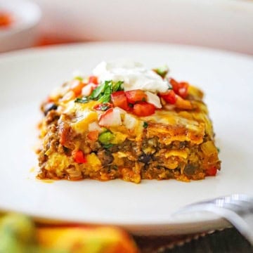 A close-up view of a square piece of a beef enchilada casserole topped with pico de gallo and a dollop of sour cream.