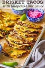 A sheet pan filled with two rows of baked brisket tacos with a colorful bowl filled with pickled red onions in one corner and another bowl filled with jalapeño-lime sauce in the opposite corner.